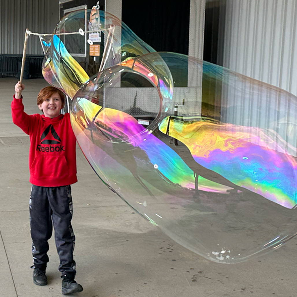 Child holding a BubbleStix wand beneath a large rainbow-colored bubble